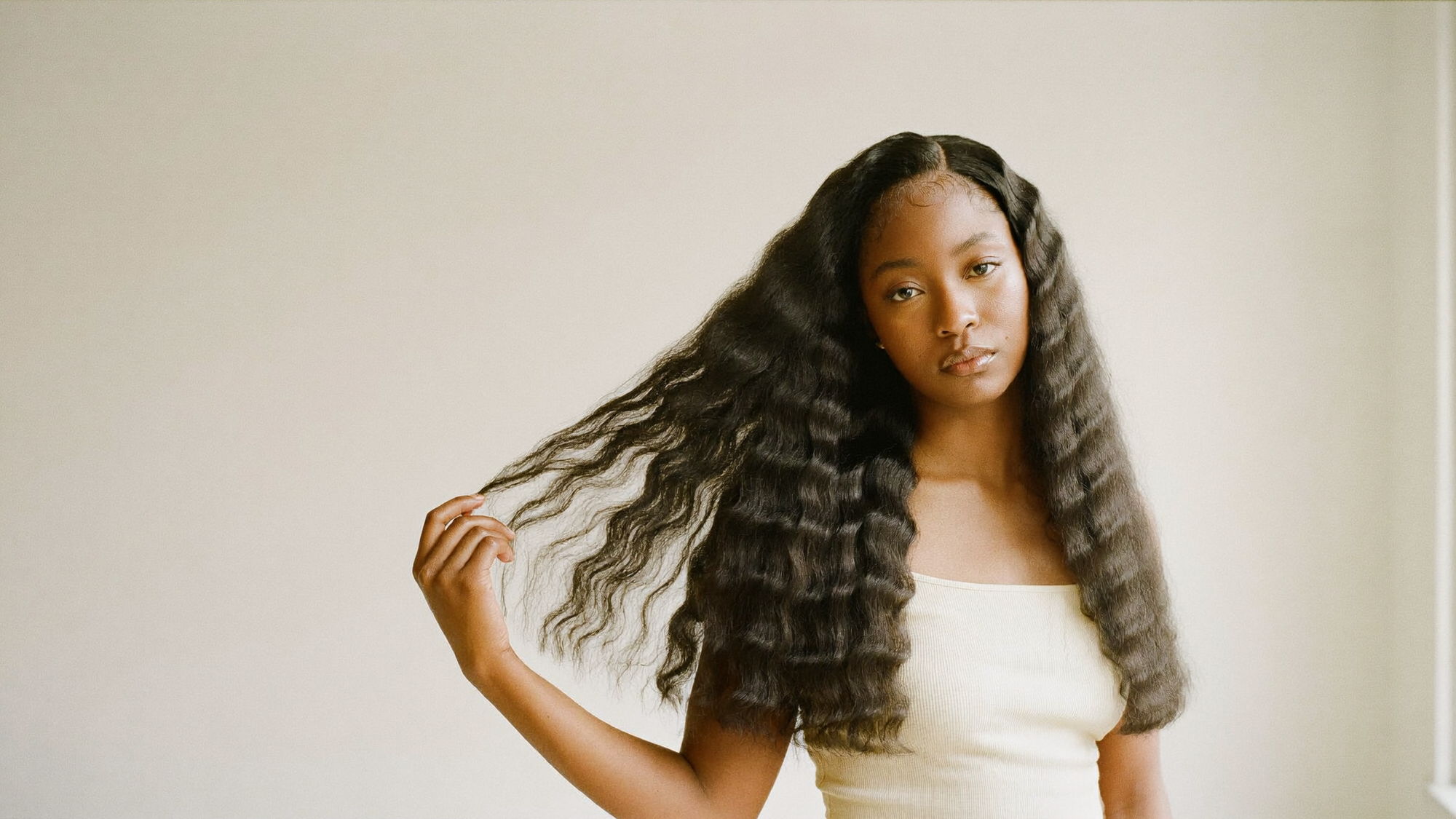 Model wearing a long wavy lace front unit, photographed against a warm cream backdrop